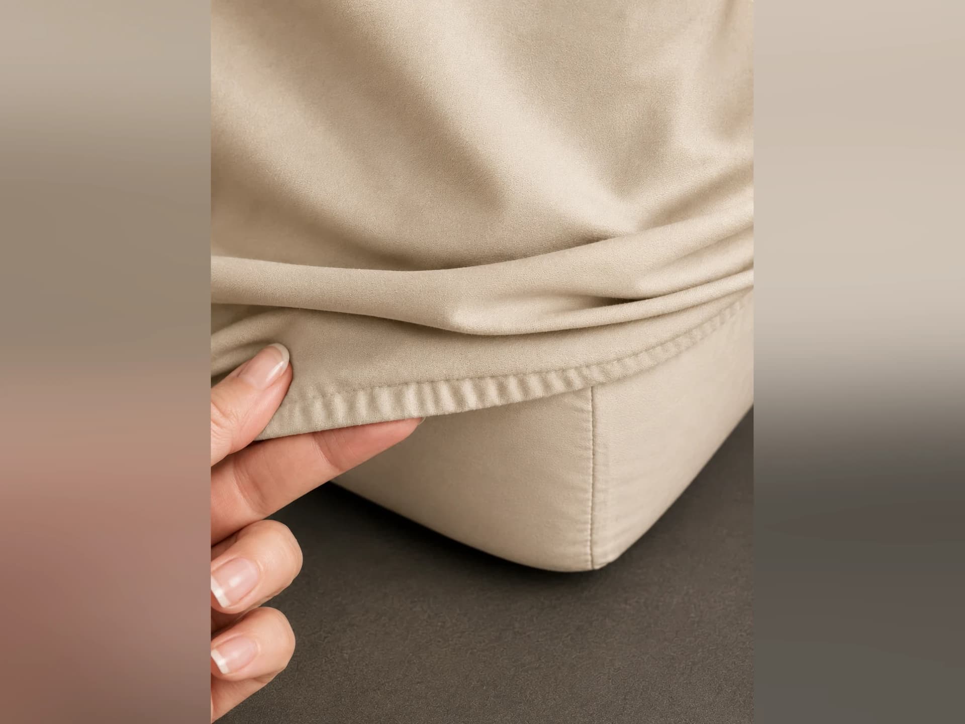 A hand lifting the elasticized hem of the fitted base at a mattress corner, showing the depth of the pocket